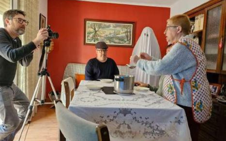 Camerògarf gravant una escena en la que es veu una taula amb cadires, una dona aixecada al costat i un home al fons de la imatge assegut a un sofà amb un fantasma al costat