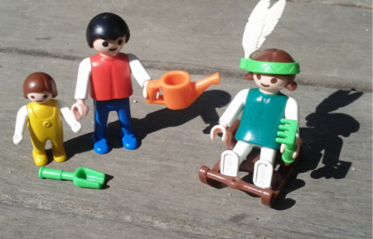 Tres ninots de playmobil sobre una estructura de fusta. Un d'ells va amb una ploma al cap com si fos un indi, l'altre porta una regadora a la mà i tercera figura petita té una pala al seu davant 