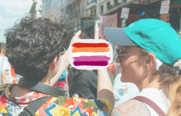 Dues noies al carrer. Un d'elles treu una foto amb mòbil. Entre elles, la bandera lèsbica